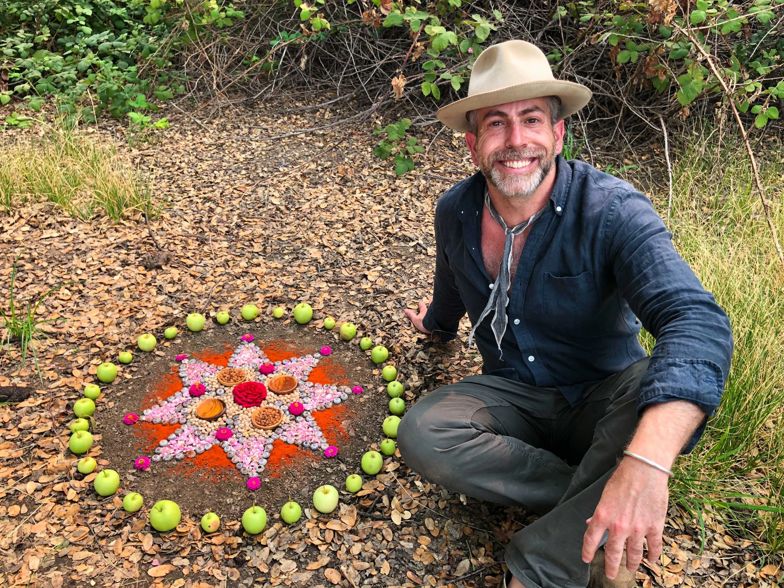 Day Schildkret kneeling beside a colorful flower-and-fruit nature altar in the woods